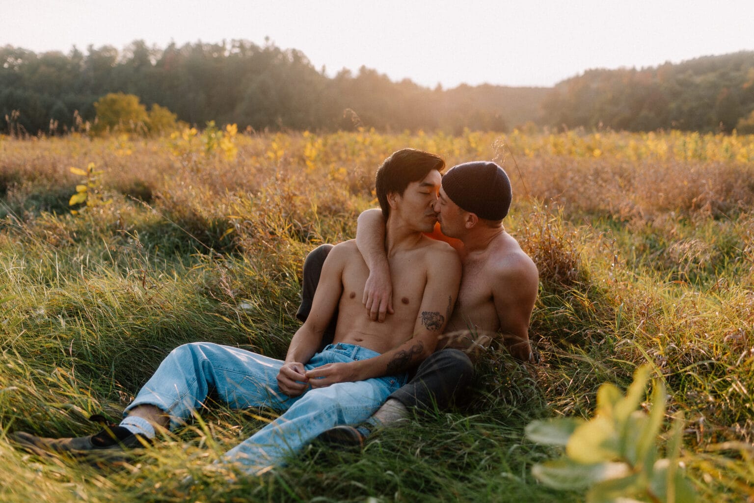 Gay couple siting in a field at sunset kissing each other.