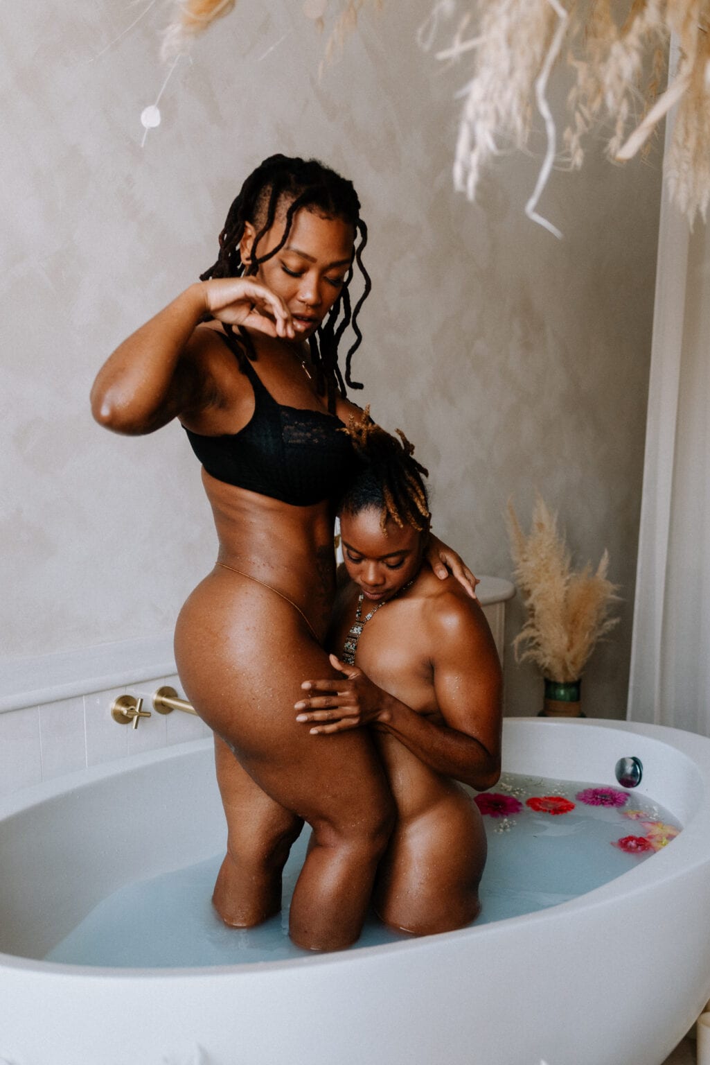 Lesbian couple taking a bath together in a bath of milk and flowers in natural light. 